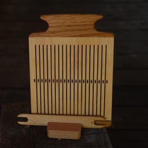 Op de afbeelding: Een houten weefgetouw met een lichtbruine afwerking. Het weefgetouw heeft verticale sleuven en een gebogen bovenkant. Een houten shuttle en een klein houten blok zijn inbegrepen. Het weefgetouw is ontworpen voor textielambachten en weefprojecten.