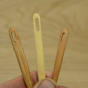 May include: Three wooden yarn needles in varying shades of light brown and tan are displayed. The needles have elongated eyelets for threading yarn and are held against a light brown wooden background. The needles are for crafts.