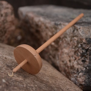 Può includere: Una ruota da filatura in legno con un fuso e un gancio. La ruota è appoggiata su una roccia.