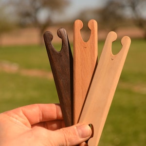 Op de afbeelding: Drie houten breinaalden in verschillende bruintinten. De naalden worden in een hand gehouden tegen een wazige achtergrond van groen gras.