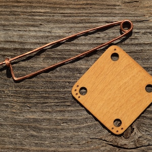 May include: A copper-coloured safety pin and a light brown wooden square with rounded corners and four holes. The wooden piece has smaller decorative holes around the larger ones. The objects rest on a weathered, grey wooden surface.