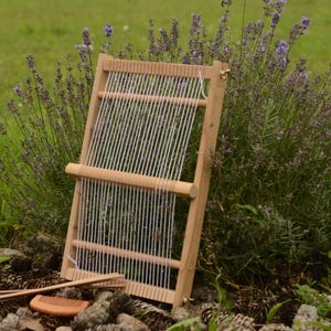 May include: A wooden weaving loom with white threads stretched across it, set against a backdrop of green grass and purple flowers. A comb and weaving tools rest at the base of the loom, suggesting a craft or hobby.