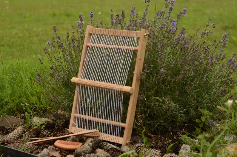 May include: A wooden weaving loom with white threads stretched across it, set against a backdrop of green grass and purple flowers. A comb and weaving tools rest at the base of the loom, suggesting a craft or hobby.