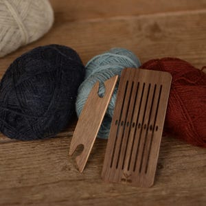 Peut inclure: Une navette de tissage en bois et un peigne en bois rectangulaire avec des fentes verticales, accompagnés de quatre pelotes de laine crème, bleu foncé, bleu clair et rouge profond. Les outils sont en bois lisse avec des découpes précises.