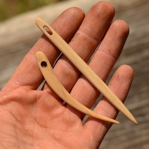 Può includere: Due aghi di legno per cucire a mano, uno dritto e uno curvo, tenuti in una mano.