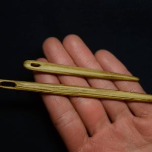 以下が含まれることがあります： 手に持たれた、目の穴のある木製の針が2本。針は薄い茶色で、濃い茶色の筋が入っている。