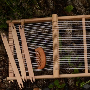 May include: A wooden weaving loom with white threads stretched across. Three wooden weaving shuttles and a wooden comb are placed near the loom. The loom is set on a bed of pine cones and greenery.