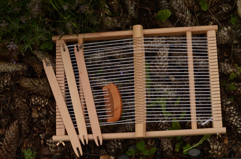 May include: A wooden weaving loom with white threads stretched across. Three wooden weaving shuttles and a wooden comb are placed near the loom. The loom is set on a bed of pine cones and greenery.