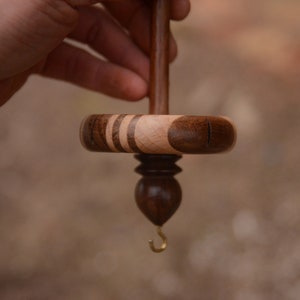 May include: A wooden spinning wheel with a brown and light wood spindle and a brass hook. The wheel is made of dark wood with light wood accents.