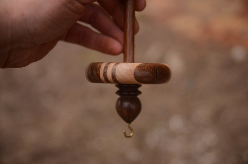 May include: A wooden spinning wheel with a brown and light wood spindle and a brass hook. The wheel is made of dark wood with light wood accents.