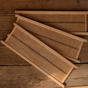 May include: Three wooden weaving looms with evenly spaced vertical slots. The looms are light brown and rectangular, with a series of parallel lines. The looms are arranged on a wooden surface.