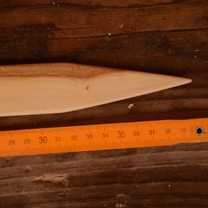 Op de afbeelding: Een lichtgekleurd houten gereedschap met een puntig uiteinde, naast een oranje vouwliniaal. De liniaal toont metingen van 63,5 cm tot 109,2 cm. Het gereedschap lijkt handgemaakt en ligt op een houten oppervlak.