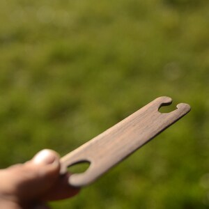 May include: A wooden weaving tool with two holes at one end and a single hole in the middle. The tool is brown and has a smooth finish.