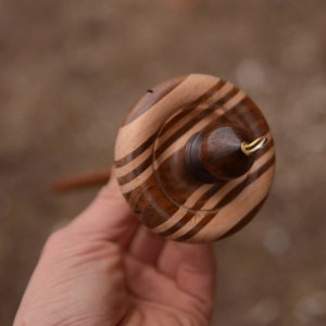 May include: A wooden spinning wheel with a brown and white striped design. The wheel has a small metal hook on the side.