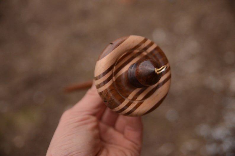 May include: A wooden spinning wheel with a brown and white striped design. The wheel has a small metal hook on the side.