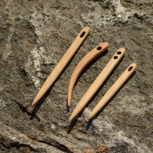 May include: Four wooden yarn needles of different shapes and sizes are displayed on a textured grey stone surface. The needles are made of light-coloured wood, with a smooth finish and large eyelets for easy threading.