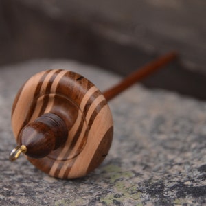 May include: A wooden spinning wheel with a brown and white striped whorl. The whorl is attached to a wooden spindle.