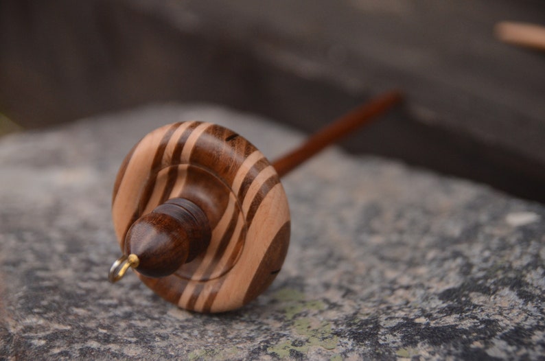 May include: A wooden spinning wheel with a brown and white striped whorl. The whorl is attached to a wooden spindle.