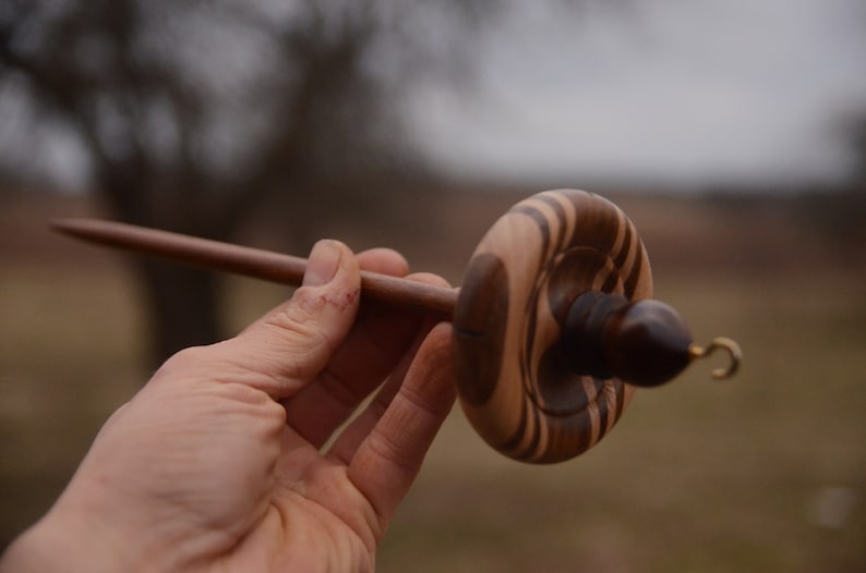 May include: A wooden spinning wheel with a brown and black striped design. The wheel has a long spindle and a hook on the end.