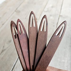 May include: Five wooden knitting needles with a dark brown finish and a light brown centre. The needles are held in a hand, fanned out in a radial pattern.