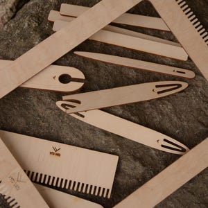Könnte beinhalten: Eine Sammlung unfertiger Webwerkzeuge aus Holz, darunter ein Rahmen, Schiffchen und ein Kamm. Die Werkzeuge sind auf einer strukturierten, grauen Oberfläche angeordnet. Die Werkzeuge sind aus hellem Holz gefertigt.