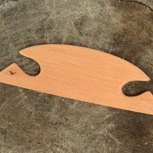 May include: A light-colored wooden tool with a unique shape, featuring two curved cutouts and a pointed end. The tool has a smooth surface and a small, dark marking near the pointed end. The tool is resting on a weathered, textured wooden surface.