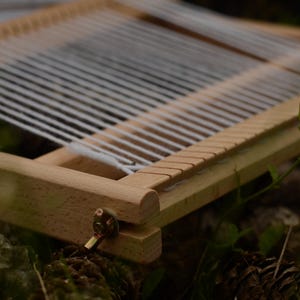 May include: A small, wooden weaving loom with white threads stretched across. The loom is rectangular with a series of notches along one edge. The wood is light-colored, and the loom is set against a blurred natural background.