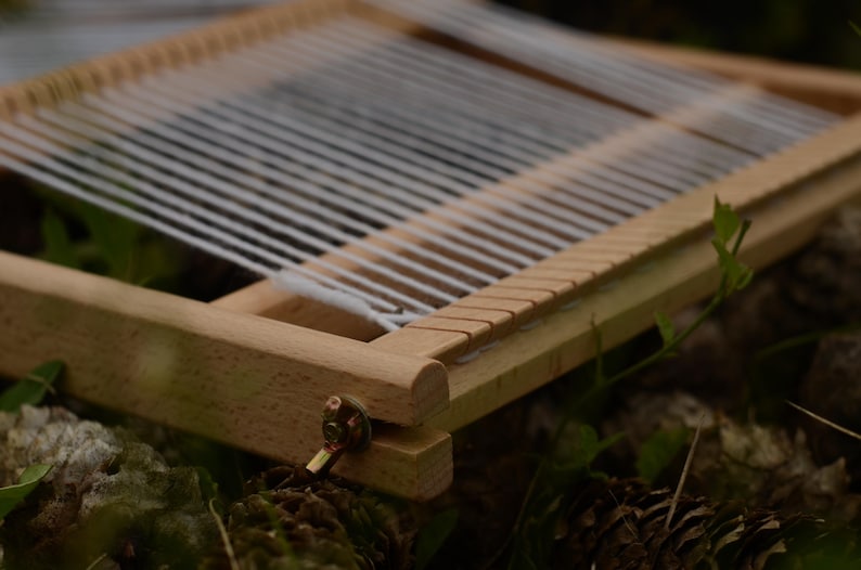 May include: A small, wooden weaving loom with white threads stretched across. The loom is rectangular with a series of notches along one edge. The wood is light-colored, and the loom is set against a blurred natural background.