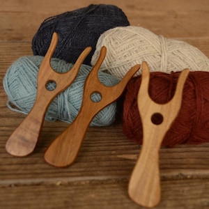 Puede incluir: Tres horquillas de lucet de madera con una parte superior en forma de Y y un orificio circular central, dispuestas frente a cuatro ovillos de lana en azul marino, crema, azul claro y rojo oscuro. Las herramientas tienen un acabado pulido.