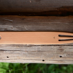 May include: A wooden knitting needle with a pointed end and a loop at the other end. The needle is lying on a wooden surface.