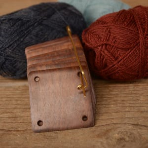 May include: A stack of square, dark brown wooden tags with four circular holes in the corners, held together by a thin gold-toned metal pin, positioned between a ball of dark gray yarn and a ball of deep red yarn.
