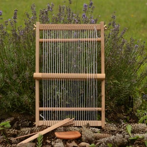 May include: A wooden weaving loom with white threads. The loom is rectangular and rests on pine cones. Two wooden tools and a small, round, orange tool are in front. Lavender bushes are in the background. The scene is outdoors.