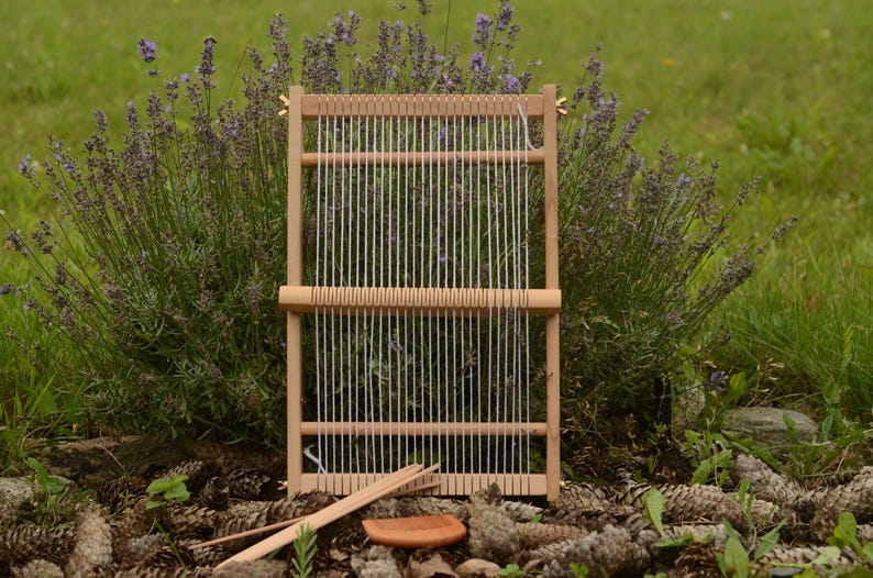 May include: A wooden weaving loom with white threads strung vertically. The loom is rectangular and sits on a bed of pine cones. Two wooden tools and a small, round, orange tool are in front of the loom. Lavender bushes are in the background.