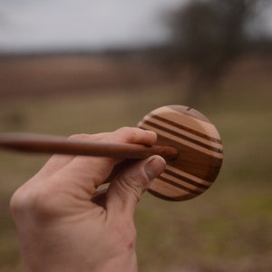 May include: A wooden spinning wheel with a brown and light wood striped design. The wheel is held in a hand.