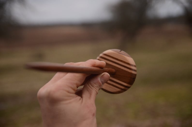 May include: A wooden spinning wheel with a brown and light wood striped design. The wheel is held in a hand.