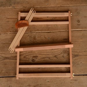 May include: A wooden weaving loom with a comb and two weaving forks. The loom is rectangular with horizontal and vertical wooden supports. The comb and forks are light brown. The loom is on a wooden surface.