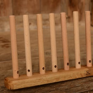 May include: A wooden plate rack with vertical dowels. The rack is made of light-colored wood and has a rectangular base. The dowels are evenly spaced and have small holes near the base. The rack is designed to hold plates upright.