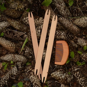 May include: Three wooden weaving shuttles and a small wooden comb rest on a bed of pine cones and green foliage. The shuttles are light brown and have a forked end. The comb is also light brown with evenly spaced teeth.