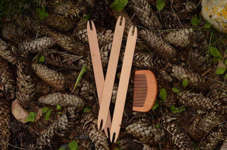 May include: Three wooden weaving shuttles and a small wooden comb rest on a bed of pine cones and green foliage. The shuttles are light brown and have a forked end. The comb is also light brown with evenly spaced teeth.