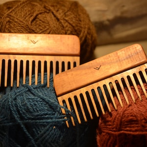 May include: Two wooden combs with a V-shaped logo on the handle. The combs are lying on top of balls of yarn in various colors, including brown, green, and blue.