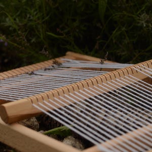 May include: A wooden weaving loom with white threads stretched across. The loom is made of light-colored wood and is set against a backdrop of green foliage. The threads are evenly spaced, ready for weaving.