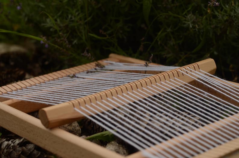 May include: A wooden weaving loom with white threads stretched across. The loom is made of light-colored wood and is set against a backdrop of green foliage. The threads are evenly spaced, ready for weaving.