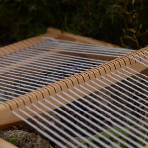 May include: A wooden weaving loom with white yarn stretched across. The loom is made of light-colored wood and has evenly spaced notches along the top and bottom edges to hold the yarn in place. The background is blurred.