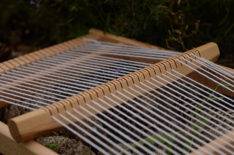May include: A wooden weaving loom with white yarn stretched across. The loom is made of light-colored wood and has evenly spaced notches along the top and bottom edges to hold the yarn in place. The background is blurred.