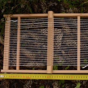May include: A wooden weaving loom with white threads stretched across. A yellow measuring tape is placed below the loom, showing measurements in inches. The loom is set against a background of pine cones and greenery.