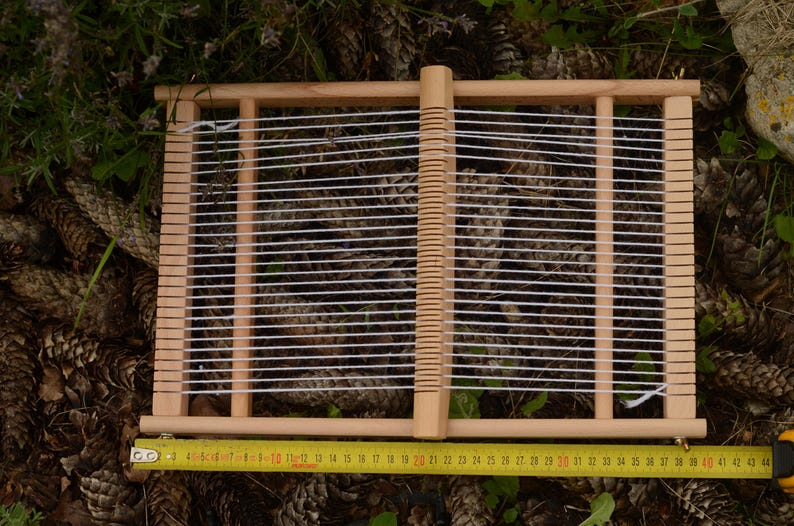 May include: A wooden weaving loom with white threads stretched across. A yellow measuring tape is placed below the loom, showing measurements in inches. The loom is set against a background of pine cones and greenery.