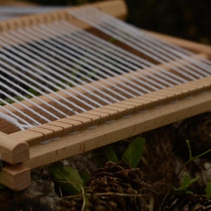 May include: A small, wooden weaving loom with white threads strung across. The loom is rectangular with a series of notches along one edge. The wood is light-colored, and the loom is set against a blurred background of greenery and pine cones.