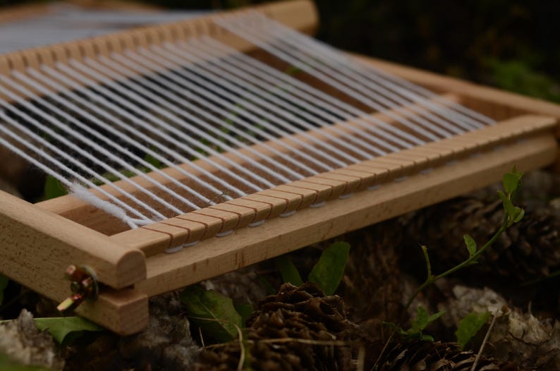 May include: A small, wooden weaving loom with white threads strung across. The loom is rectangular with a series of notches along one edge. The wood is light-colored, and the loom is set against a blurred background of greenery and pine cones.