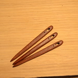 Könnte beinhalten: Drei hölzerne Faden-Nadeln mit einem kleinen Loch oben. Die Nadeln sind braun und haben eine glatte Oberfläche.