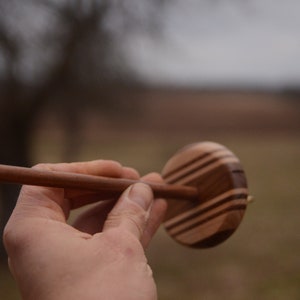 May include: A wooden spinning wheel with a brown and black striped design. The wheel is held in a person's hand.